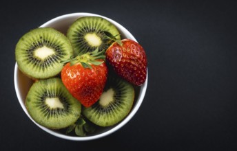 Sliced kiwis and strawberries in a white bowl with a black background