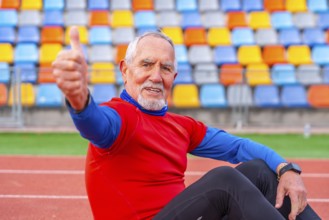 Proud senior man gesturing with thumbs up sitting on a running track after running
