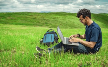 Close up of man sitting on green grass working with his laptop. Smiling freelancer guy working from