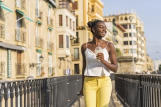 African young woman carrying luggage and a take away beverage along a bridge in the street