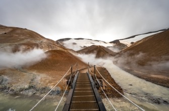 Bridge over steaming stream between colourful rhyolite mountains with snowfields, Hveradalir