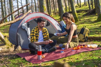 Hikers playing cards near their tent in the forest, enjoying a camping trip with fruits and snacks