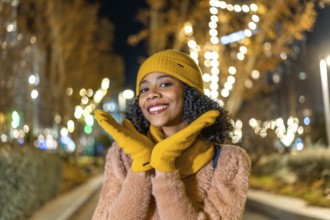 Portrait of a cheerful young woman wearing yellow woolen gloves and hat enjoying christmas lights