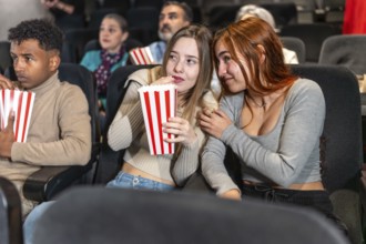 Two friends are whispering and eating popcorn while watching a movie at the cinema, surrounded by