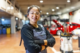 Confident young female mechanic wearing safety glasses and earplugs, smiling with crossed arms in a