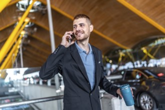 Young businessman smiling while talking on phone, holding his luggage and coffee thermos, at the