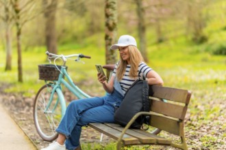 Young woman is using her smartphone while sitting on a bench in a park, with her bicycle parked