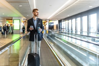 Businessman is walking on moving walkway at airport while using smartphone and holding coffee