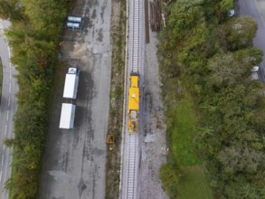 Top view of a yellow construction machine on a railway track next to a road and a storage yard,