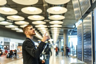 Businessman consulting his smartphone and checking flight information displayed on screens at the
