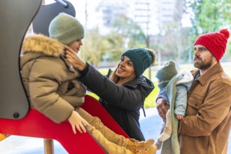 Smiling mother helping her son going down the slide, while father holding baby, enjoying a winter