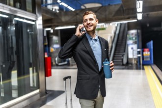 Young businessman talking on mobile phone and holding reusable coffee cup while waiting for subway