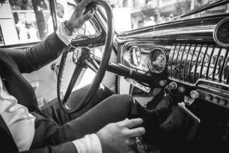 A man in a suit is driving a vintage car. The car is black and white. The man is wearing a watch