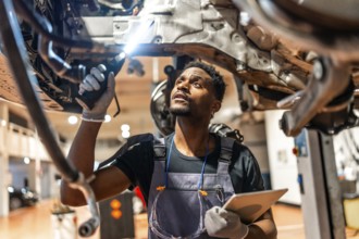 Young mechanic inspecting car undercarriage using flashlight and holding tablet in a modern auto