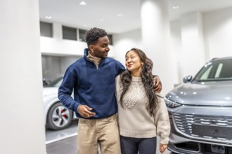 Happy multiethnic couple buying a new electric car at a dealership, hugging each other and smiling