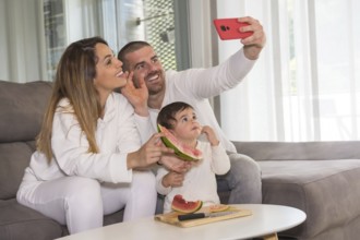 Family of three waving at camera taking a selfie while eating watermelon sitting on the sofa at