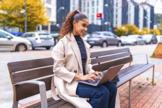 African american elegant businesswoman sitting on a bench using laptop in the city in autumn