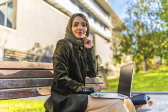 Young Muslim businesswoman smiling at camera while using laptop sitting on park bench