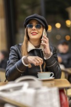 Fashionable woman wearing sunglasses and hat stirring coffee while talking on mobile phone at a