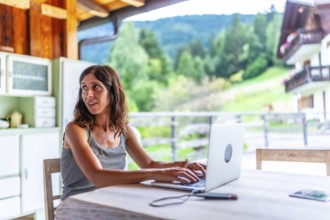 Freelancer woman working remotely on vacation with a laptop in a mountain chalet, enjoying the view