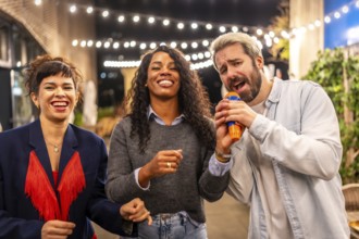 Three diverse friends singing karaoke and laughing at night party in the roof terrace of a bar