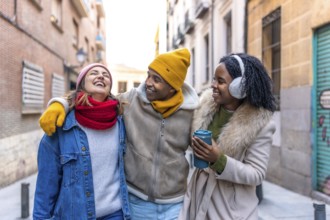 Three happy multi ethnic friends are walking embracing in the city during winter, laughing and