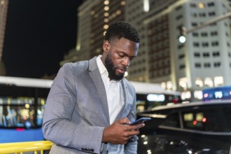 Young businessman using smartphone on a city street at night, with office buildings and traffic in