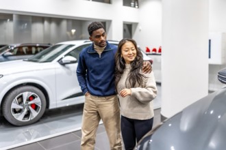 Happy multiethnic couple buying a new electric car at the dealership, embracing and choosing their