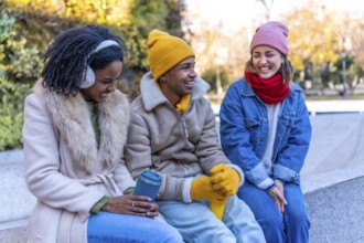 Three young multi ethnic friends wearing winter clothes chatting and laughing, sitting on a bench