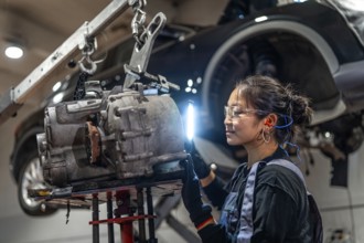 Young woman working as mechanic inspecting gearbox illuminated with flashlight in car workshop