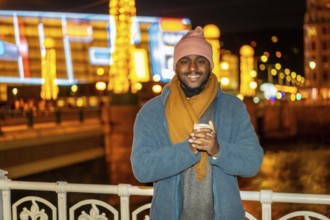 Smiling african man holding a warm cup of coffee in the city in a Christmas season night