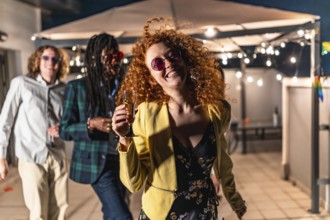 Happy redhead woman dancing passionately at night terrace party with multiracial friends