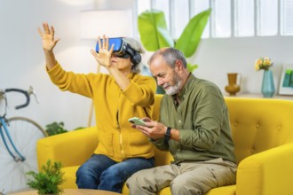 Senior woman using virtual reality googles next to her husband sitting on the sofa using mobile