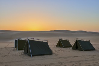 Four tents stand in a vast desert at dusk, surrounded by sand dunes under an orange-coloured sky,