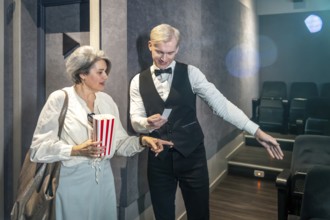 Cinema usher showing assigned seat to elegant mature woman holding popcorn bucket while entering