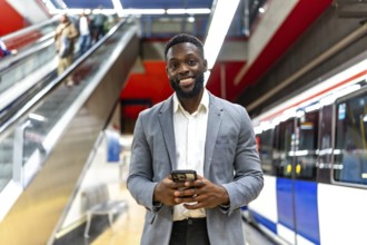 Young black businessman using smartphone while waiting for subway train in station, with escalator