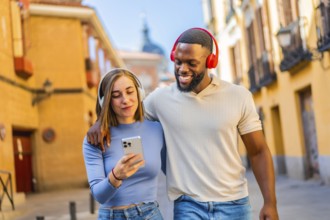 Multiracial happy couple walking in the city using mobile phone and headphones