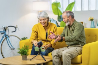 Senior couple smiling during video call using smartphone sitting in a yellow modern sofa at home