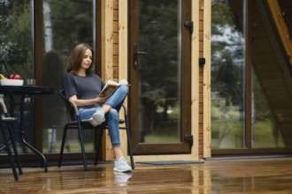 Cute woman sits on terrace of her forest house and reads the book
