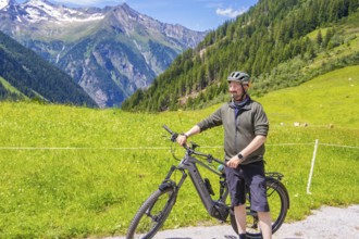 Man in sportswear standing next to his bicycle on a green meadow, surrounded by mountains and blue