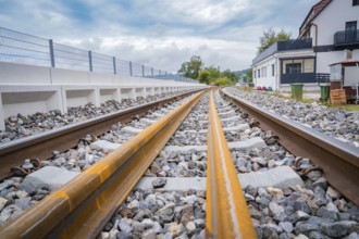 A railway track runs through a landscape with buildings in the background, under a cloudy sky,