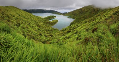 Natural landscape with a green slope and a lake surrounded by hills and a cloudy sky, crater lake