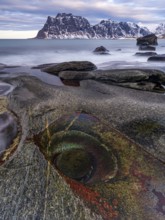 Rocks on the beach, rock formation Eye of Utakleiv, or Dragon's Eye, snow-capped mountains behind,