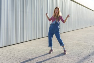 Blonde dancer performing urban dance moves on a paved area against a corrugated metal wall,