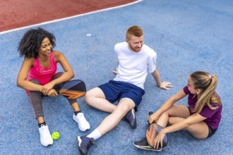 Multi-ethnic friends sitting on the ground relaxed and talking in a pickelball court