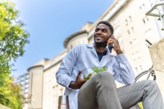 African young doctor talking to the mobile while eating lunch outside the hospital