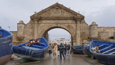 People walking through a stone arch, flanked by blue fishing boats, under a cloudy sky, Essaouira,