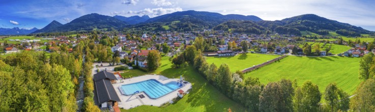 Panoramic view of a village with a swimming pool, surrounded by green meadows and forests, against