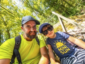 Father and son taking a selfie while hiking in a lush green forest, enjoying their summer vacation