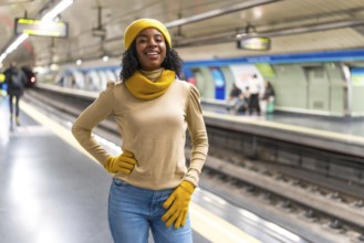 Smiling young woman wearing yellow wool cap and gloves is waiting for subway train arrival at